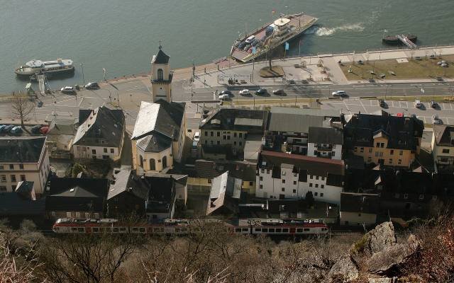 A red regional train passing along the riverfront of Sankt Goarshausen, seen from above